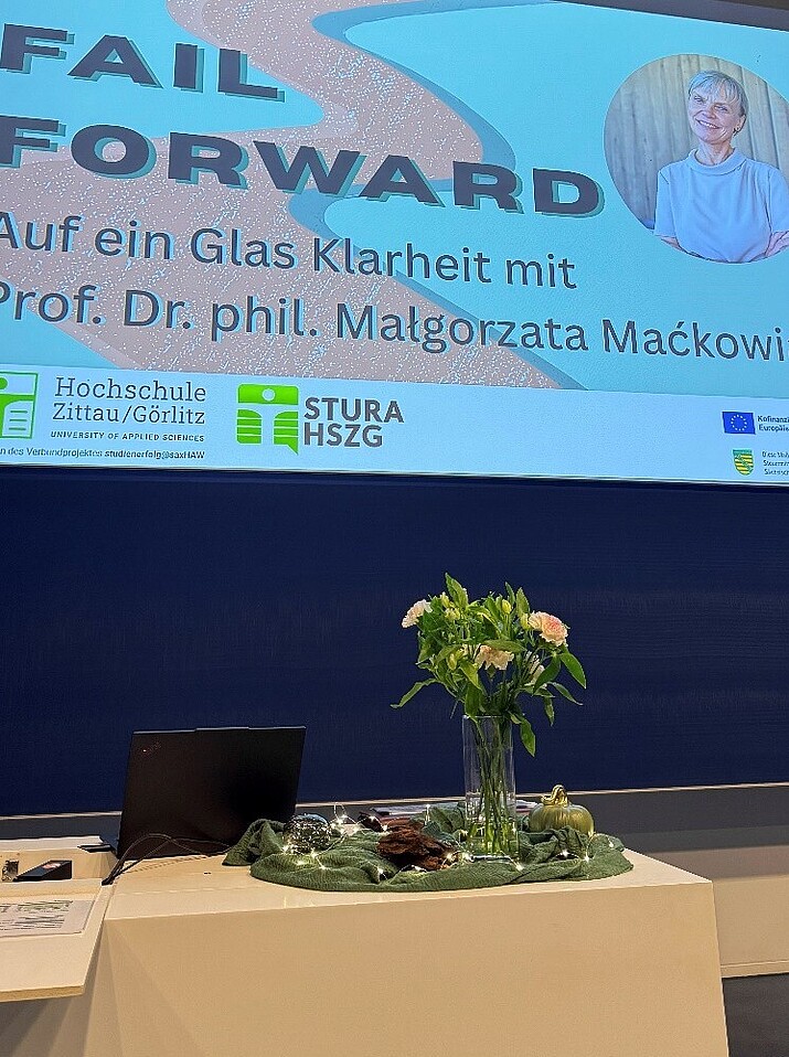 Eine Blumenstrauß steht neben einem geöffneten Laptop auf einem Rednerpult, im Hintergrund erscheint eine Beamerprojektion.