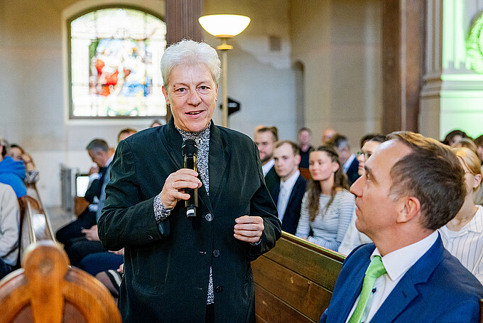 Ellen Leuschner steht mit einem Mikrofon zwischen den Kirchenbänken und spricht zum Publikum.