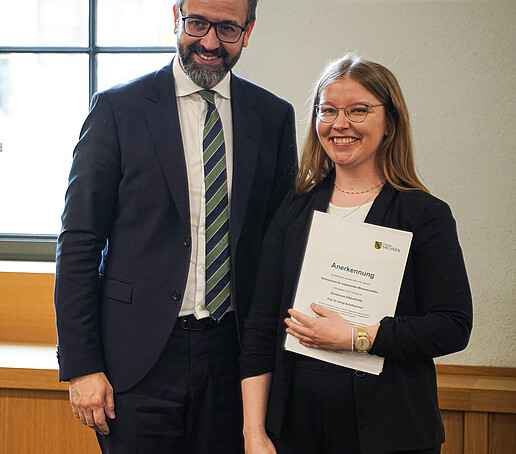 Ein Mann und eine Frau stehen nebeneinander, beide lächeln in die Kamera. Die Frau hält eine Urkunde mit dem Logo des Freistaates Sachsen.