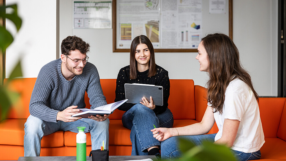 Drei Studierende sitzen auf einem orangenen Sofa und unterhalten sich.