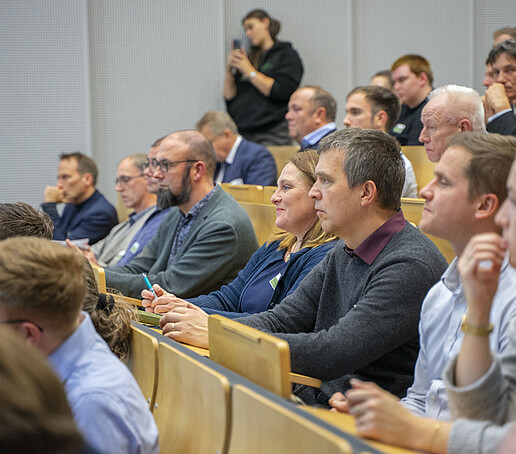 Die Sitzreihen sind mit Publikum besetzt, dass den Blick nach vorn richtet.