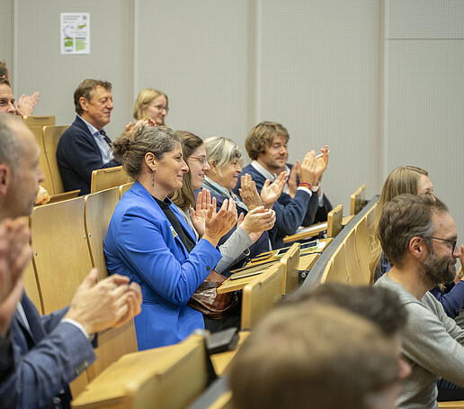 Das Publikum sitzt applaudierend auf den Sitzreihen.