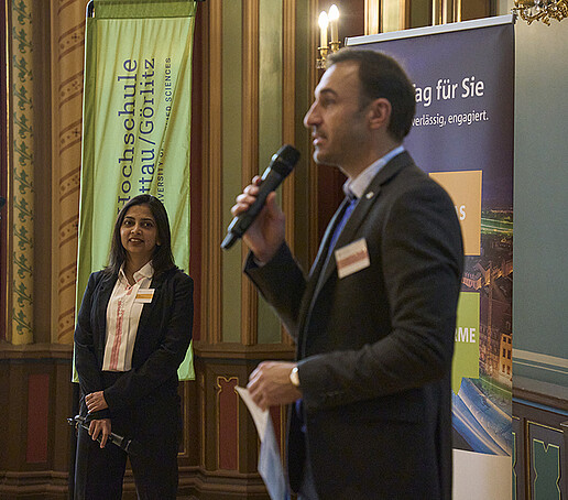 Prof. Schütte steht vor dem Publikum neben einer Teilnehmerin und spricht in ein Mikrofon.