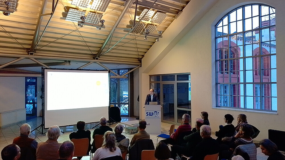 Seniorinnen und Senioren sitzen auf Sitzreihen vor einer Leinwand im E-Werk. Prof. Kunick steht hinter einem Rednerpult.