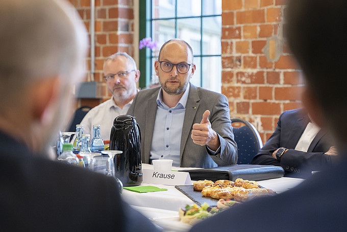 Ein Mann mit Brille spricht und gestikuliert während einer Besprechung am Tisch, weitere Teilnehmende hören zu und schauen ihn an.