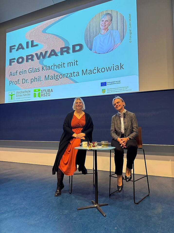 Zwei Frauen sitzen auf Hochstühlen im Vorlesungssaal vor einer Beamerprojektion.