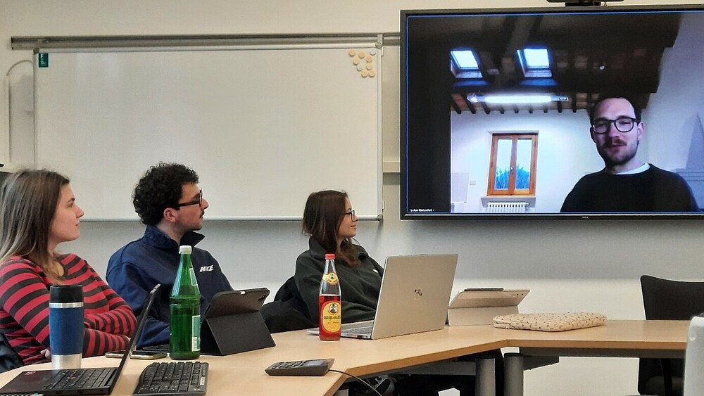 Studierende sitzen im Seminarsaal am Tisch und sehen auf einen Bildschirm auf dem der Autor Lukas Rietzschel zugeschaltet ist.