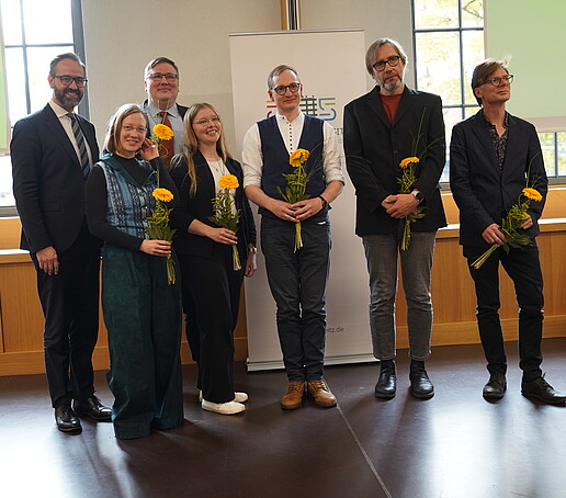 Sieben Personen stehen nebeneinander, halten Blumen in den Händen und blicken in die Kamera.