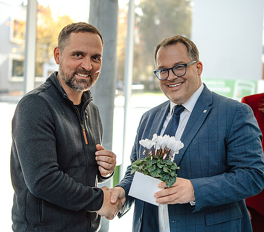 Der Rektor schüttelt einem Professor die Hand und hält ein Blumengesteck in den Händen.